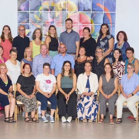 Eine Gruppe von Erwachsenen, sowohl Männer als auch Frauen, sitzen auf Stühlen und stehen in einem Raum, wahrscheinlich für ein Gruppenfoto posieren. Ein farbenfrohes Wandbild befindet sich hinter ihnen.