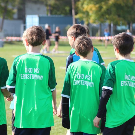 Eine Gruppe junger Jungen, die grüne Trikots mit weißem Text auf dem Rücken tragen, steht auf einem Rasenfeld. Der Text lautet 'OKO MS Ernstbrunn'. Im Hintergrund sind verschwommene Personen und Bäume zu sehen.