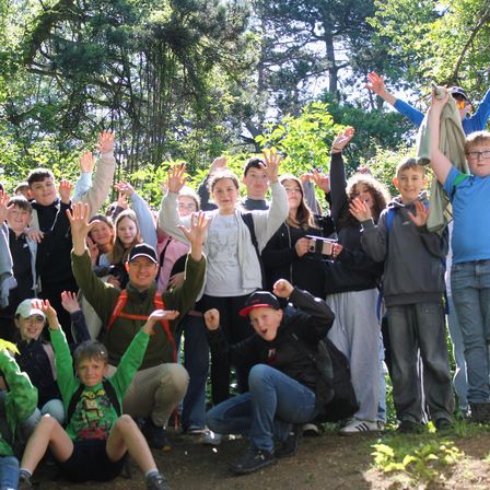 Eine Gruppe junger Menschen in einem Wald, einige stehend und andere sitzend, alle lächeln und winken zur Kamera.