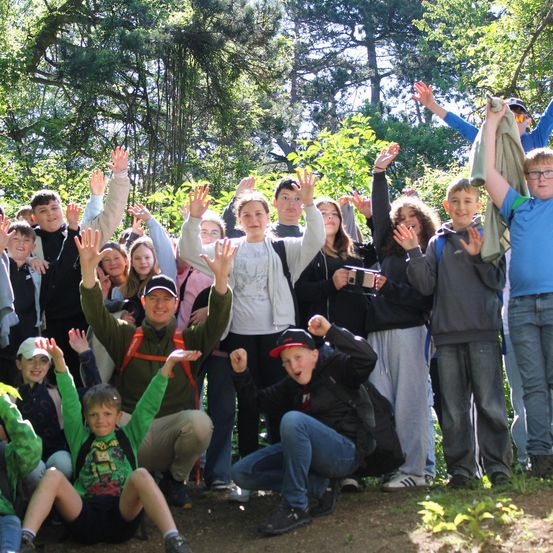Eine Gruppe junger Menschen in einem Wald, einige stehend und andere sitzend, alle lächeln und winken zur Kamera.