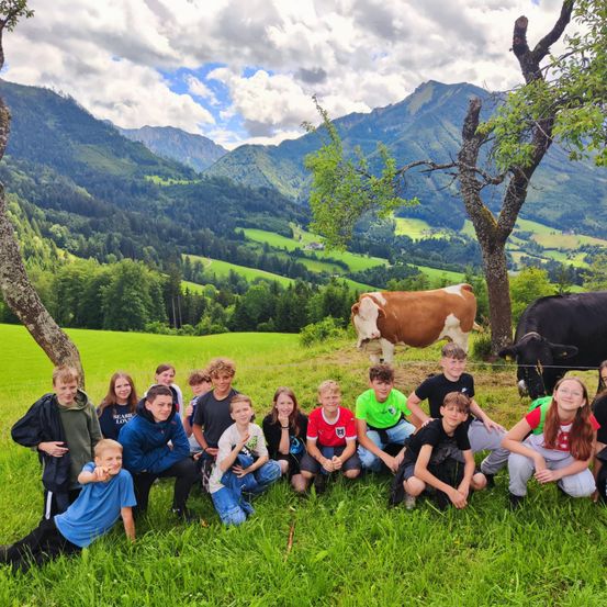 Eine Gruppe von Kindern und Jugendlichen posiert für ein Foto auf einer Wiese mit Kühen und Bergen im Hintergrund.