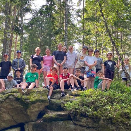 Eine Gruppe junger Menschen, sowohl Jungen als auch Mädchen, posiert für ein Foto in einer Waldumgebung, wobei einige auf Felsen sitzen und andere hinter ihnen stehen.