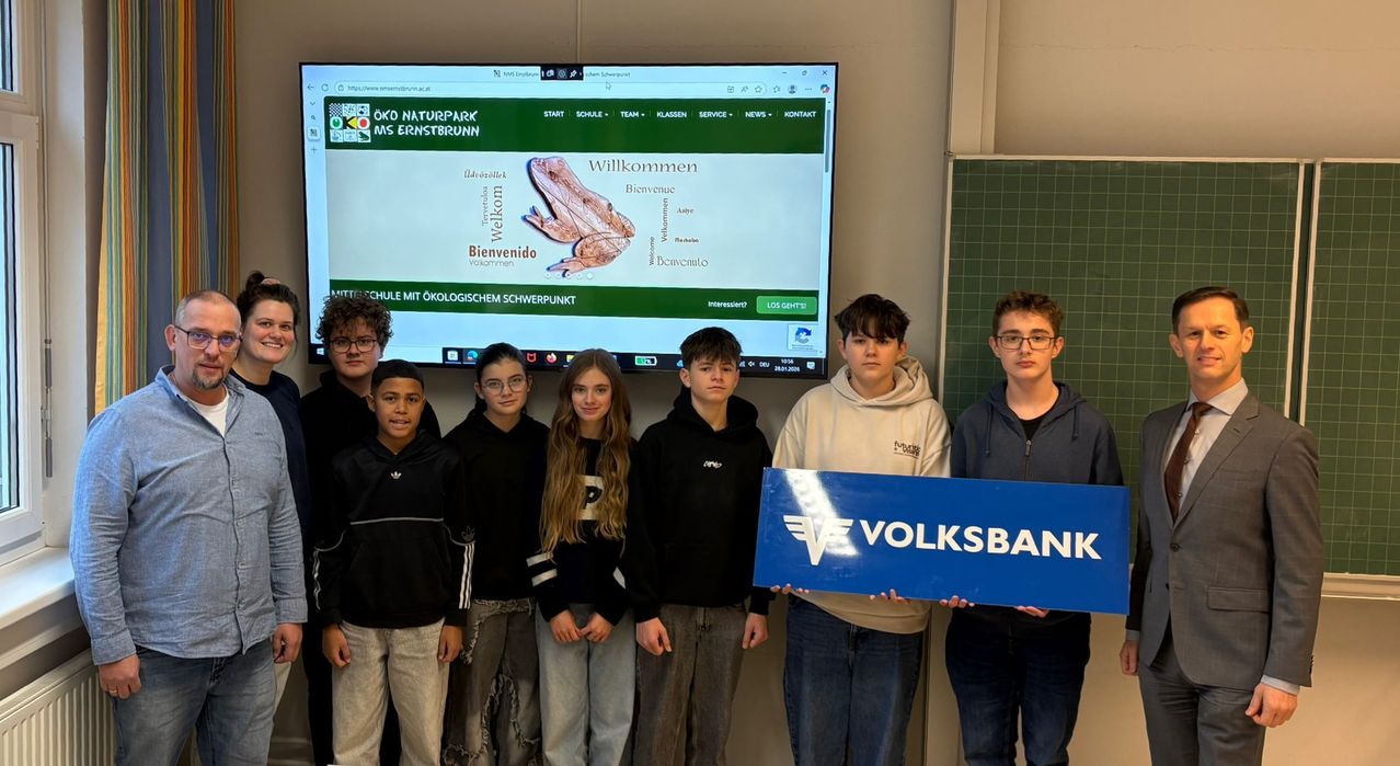 Schüler stehen in einem Klassenzimmer und halten ein blaues Schild für Volksbank. Dahinter zeigt ein großer Bildschirm die Website 'Oko Naturpark MS Ernstbrunn' an.