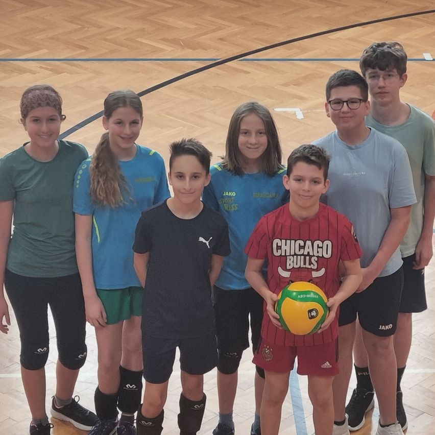Eine Gruppe von Kindern steht auf einem Basketballplatz, trägt Sportbekleidung und hält einen Volleyball. Sie lächeln und posieren für ein Foto.