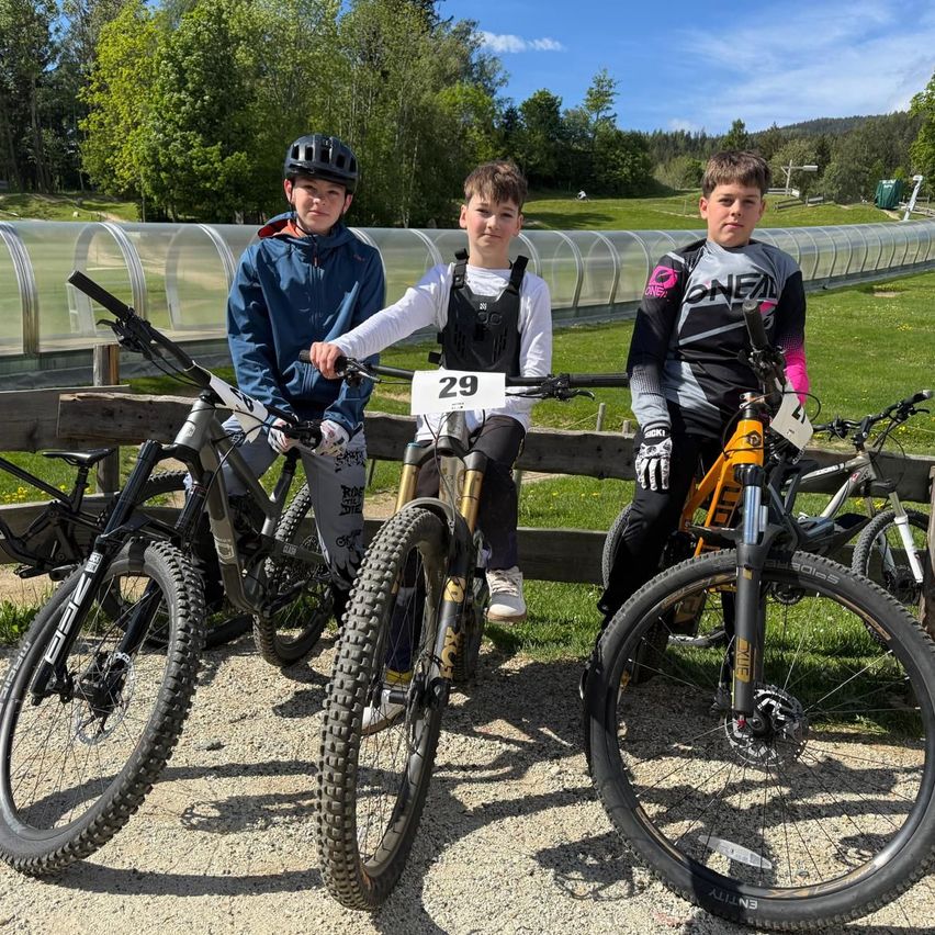 Drei junge Jungen in Fahrradtrikots mit Nummern darauf posieren mit ihren Fahrrädern für ein Foto in einem Park.