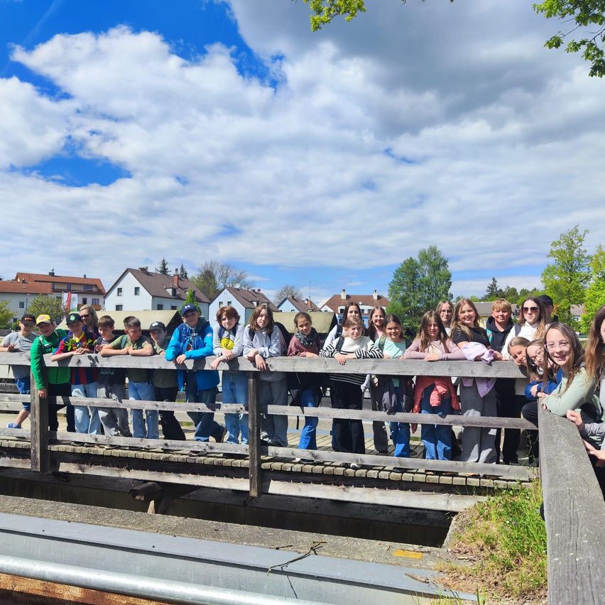 Eine Gruppe von Menschen steht auf einer Holzbrücke über einem Gewässer mit Häusern und Bäumen im Hintergrund.