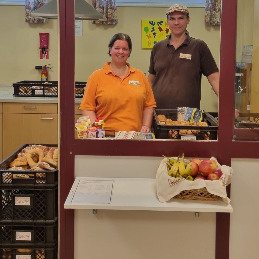 Zwei Personen stehen hinter einer Theke in einer Bäckerei. Eine Frau in einem orangefarbenen Shirt und ein Mann in einem braunen Shirt lächeln. Es gibt Körbe mit Brot, einen Korb mit Obst und eine Speisekarte auf der Theke.