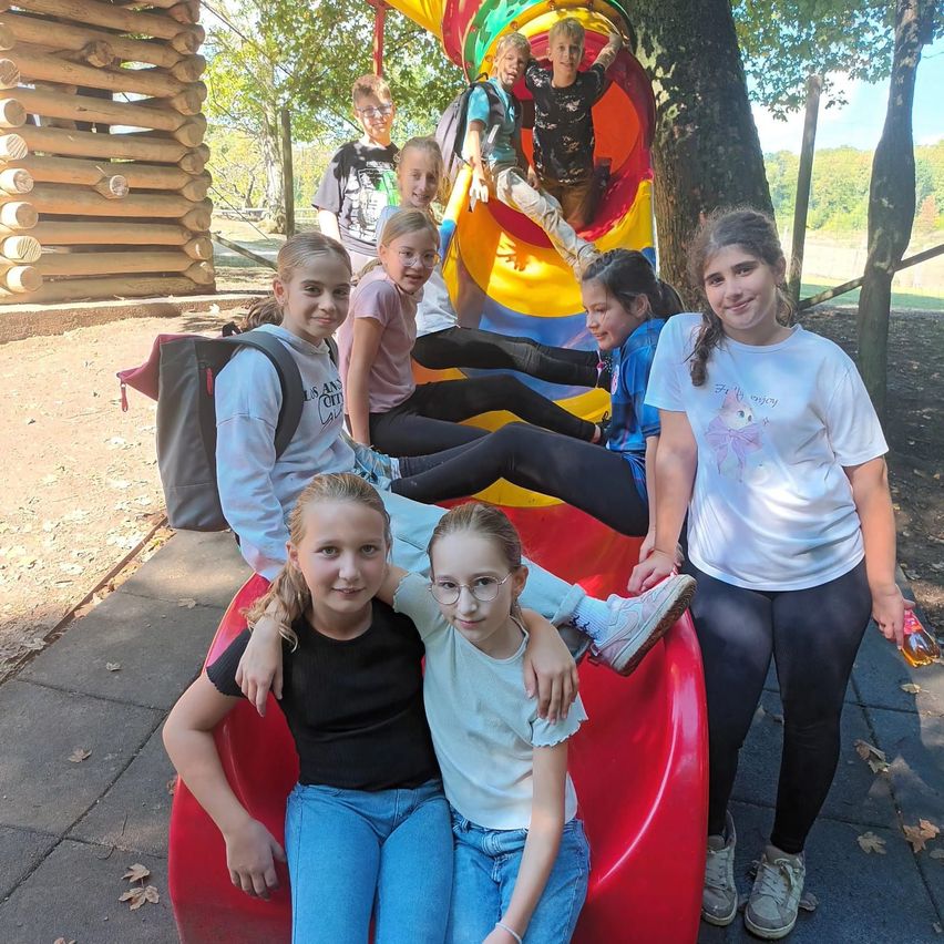 Eine Gruppe junger Mädchen und Jungen spielt auf einer bunten Rutsche in einem Park. Einige sitzen auf der Rutsche, während andere darum herumstehen.
