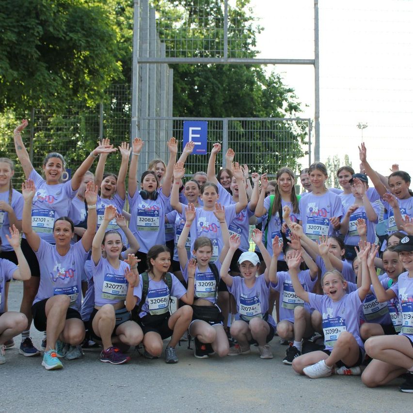 Eine Gruppe junger Mädchen in lila T-Shirts mit Nummern posiert für ein Foto bei einem Marathon-Event. Sie heben ihre Hände und lächeln, während sie vor einem Metallzaun mit einem blauen Schild stehen.