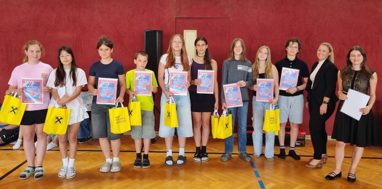 Eine Gruppe von Kindern und Jugendlichen steht zusammen, hält Auszeichnungen und gelbe Taschen mit der Aufschrift 'Wir machts möglich'. Sie posieren für ein Foto in einem Raum mit roter Wand.