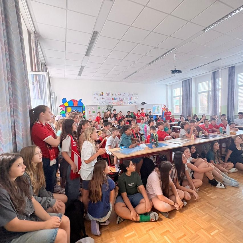 Ein Klassenzimmer voller Schüler, die um einen langen Tisch herum sitzen. Einige Schüler stehen an den Wänden und beobachten das Ereignis. Der Raum hat einen Holzboden, Vorhänge und Poster an der Wand.