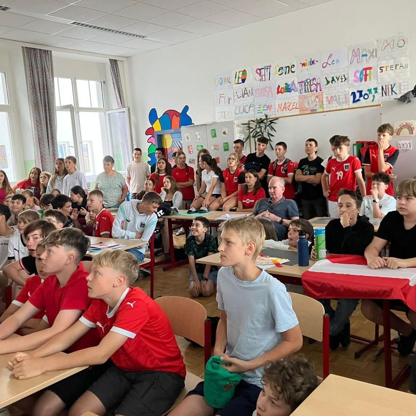 Ein Klassenzimmer mit Schülern in roten und weißen Shirts, die an Tischen sitzen. Einige tragen Turnschuhe, andere Sandalen. Ein Tisch davor hat eine rote Tischdecke. Eine farbenfrohe Wand mit Zeichnungen und Namen.