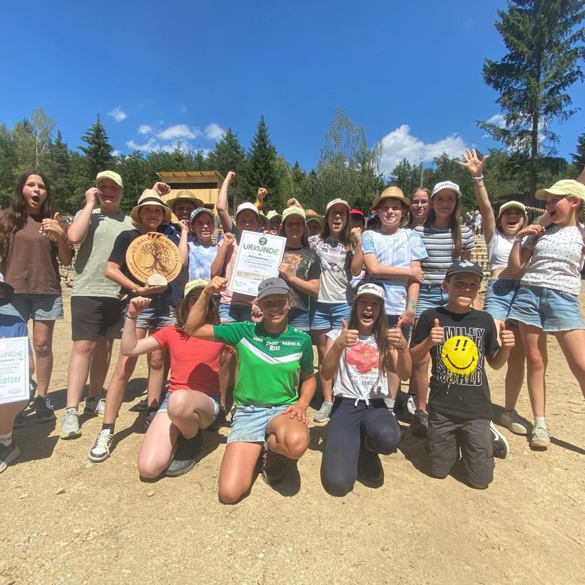Eine Gruppe von Kindern in Hüten und Turnschuhen posiert für ein Foto im Freien, einige halten Urkunden in der Hand. Die Sonne scheint, und der Boden ist mit kleinen Steinen bedeckt. Bäume und eine Holzstruktur sind im Hintergrund zu sehen.