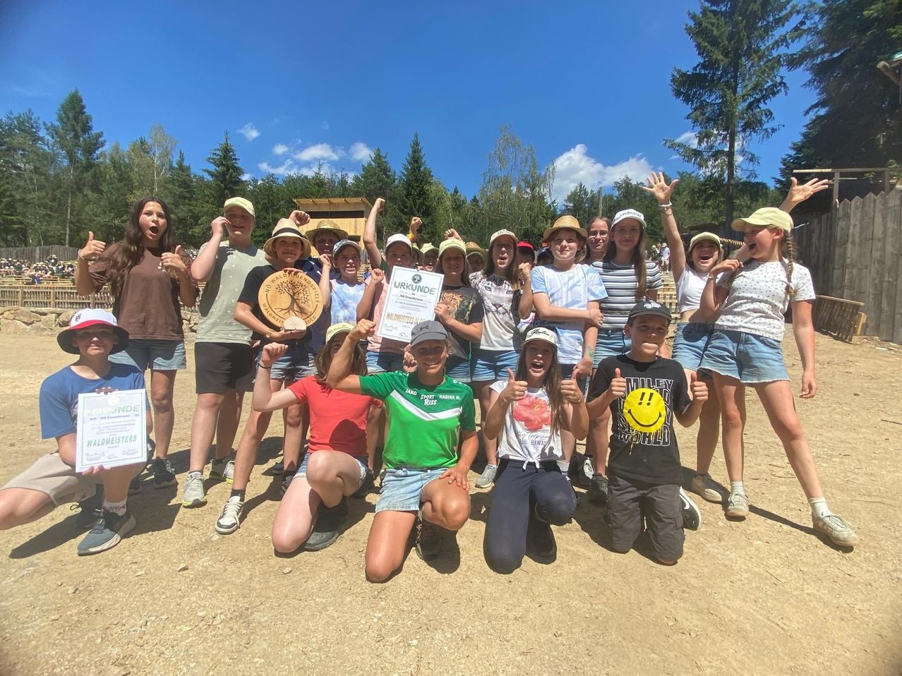 Eine Gruppe von Kindern in Hüten und Turnschuhen posiert für ein Foto im Freien, einige halten Urkunden in der Hand. Die Sonne scheint, und der Boden ist mit kleinen Steinen bedeckt. Bäume und eine Holzstruktur sind im Hintergrund zu sehen.