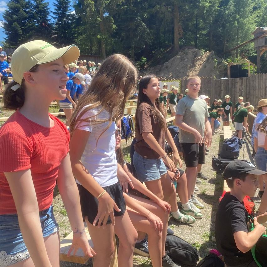 Mehrere junge Leute sitzen auf Tribünen im Freien. Ein Mädchen in einem roten T-Shirt schaut ein anderes Mädchen vor sich an, das ein weißes T-Shirt trägt. In der Nähe steht ein Junge in einem braunen T-Shirt mit verschränkten Armen.
