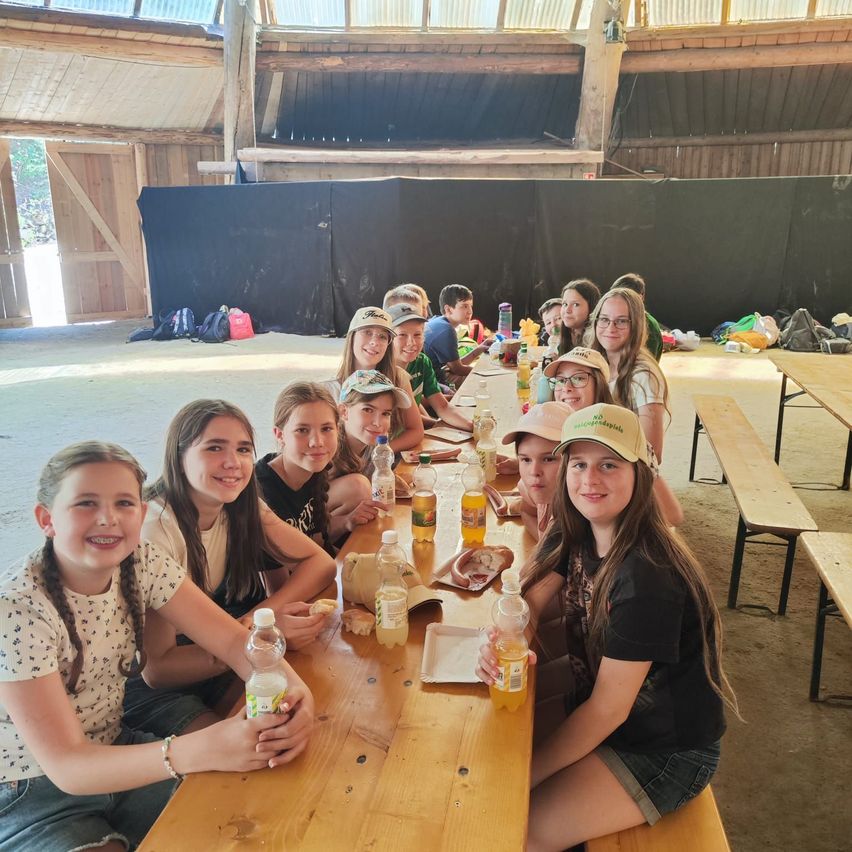 Eine Gruppe junger Mädchen und Jungen sitzt an einem langen Tisch in einer Scheune, trägt Hüte und hält Flaschen mit Saft. Sie lächeln und scheinen ihre Mahlzeit zu genießen. Der Tisch ist mit Tellern, Tassen und anderen Gegenständen gefüllt. Im Hintergrund befindet sich eine Bühne mit schwarzem Hintergrund.