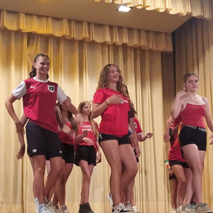 Eine Gruppe junger Frauen in roten Shirts und schwarzen Shorts tritt auf der Bühne auf, mit gelben Vorhängen im Hintergrund.