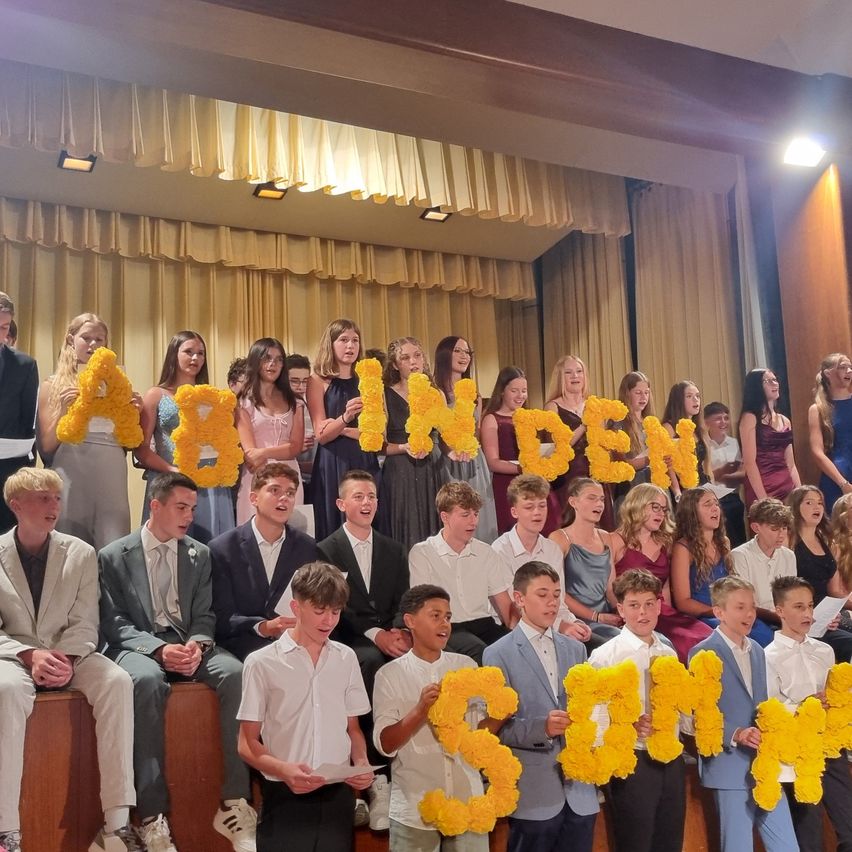 Ein Chor von Schülern, Jungen und Mädchen, steht auf einer Bühne und hält gelbe Blumen, die 'Abinden' und 'Sommer' buchstabieren.