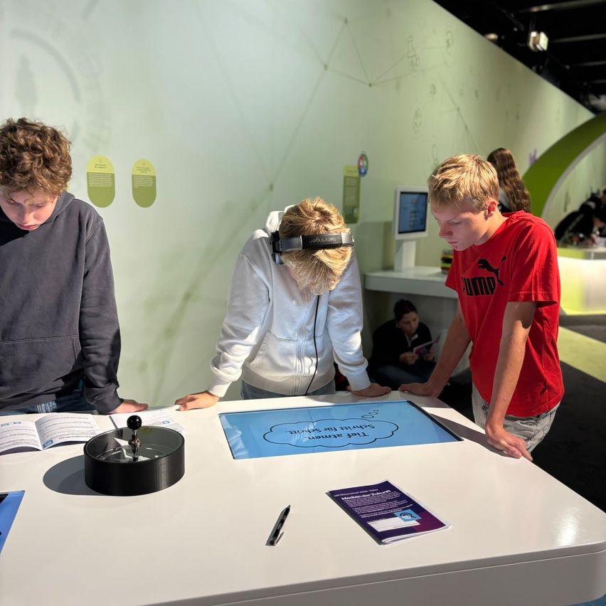 Drei junge Personen sind in einer interaktiven Ausstellung in einem Wissenschaftsmuseum beschäftigt. Eine Frau in einer weißen Kapuzenjacke und Kopfhörern zeigt auf einen digitalen Bildschirm, während zwei Jungen, einer in einem roten Shirt, zuschauen.
