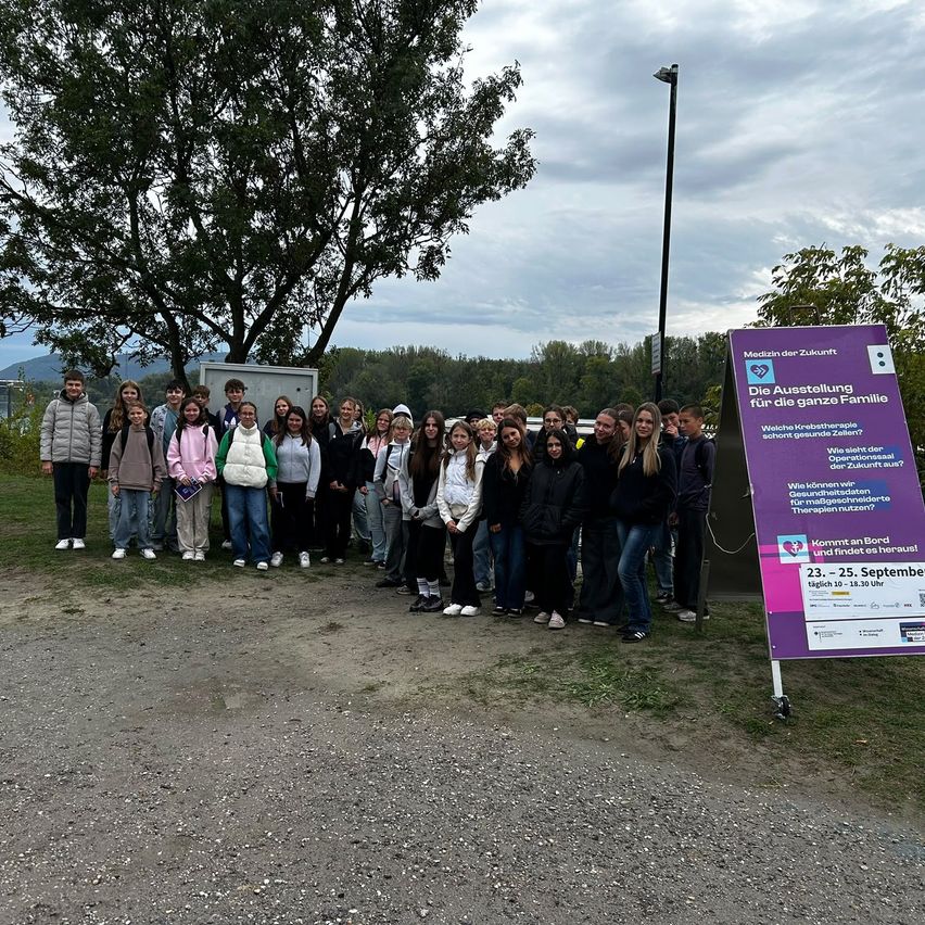 Eine Gruppe von Menschen steht vor einem violetten Banner mit Text, wahrscheinlich bei einer medizinischen Ausstellung. Bäume und ein bewölkter Himmel im Hintergrund.