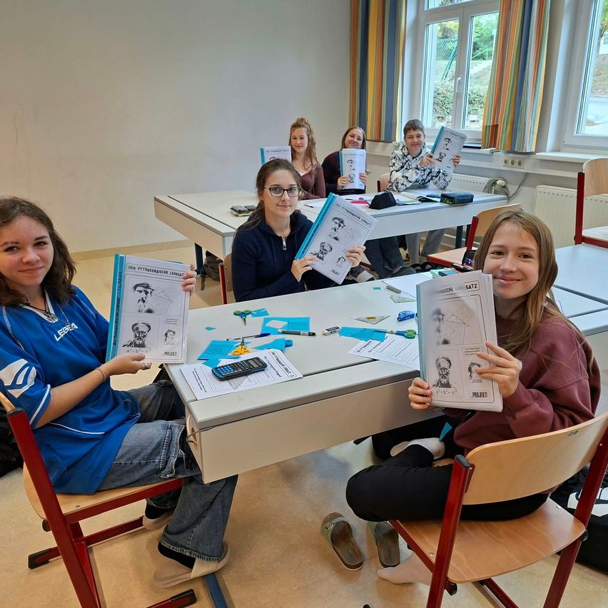 Eine Gruppe von Schülern im Klassenzimmer, jeder hält ein Buch mit einer Zeichnung eines Mannes auf dem Cover. Sie sitzen an Tischen mit verschiedenen Papieren und einem Taschenrechner.