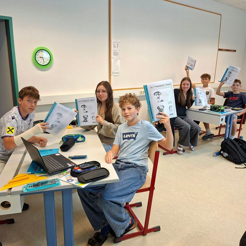Ein Klassenzimmer mit Schülern, die Bücher hochhalten und lächeln, einer mit einem verbundenen Arm, einer Uhr an der Wand und einem Laptop auf dem Schreibtisch.