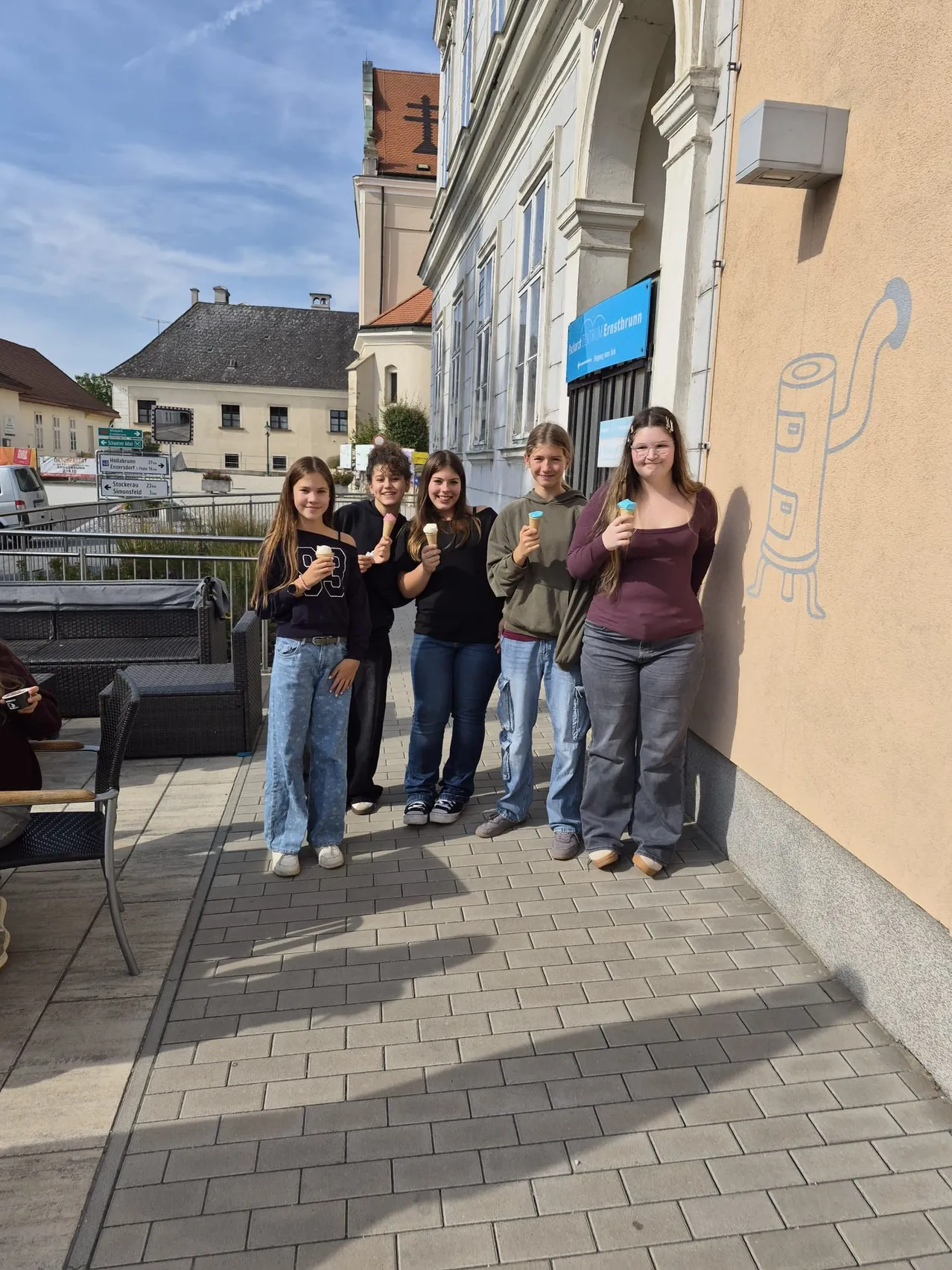 Fünf junge Frauen stehen auf einem gepflasterten Bürgersteig, jede hält einen Eisbecher. Sie stehen vor einem Gebäude mit einer Wandmalerei.