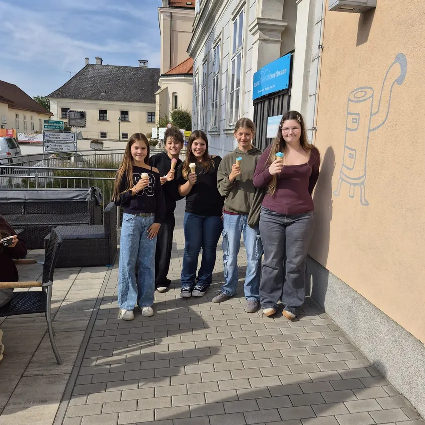 Fünf junge Frauen stehen auf einem gepflasterten Bürgersteig, jede hält einen Eisbecher. Sie stehen vor einem Gebäude mit einer Wandmalerei.