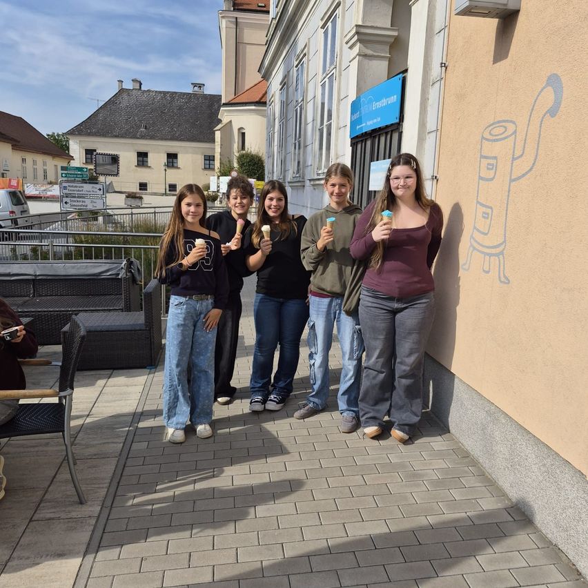 Fünf junge Frauen stehen auf einem gepflasterten Bürgersteig, jede hält einen Eisbecher. Sie stehen vor einem Gebäude mit einer Wandmalerei.