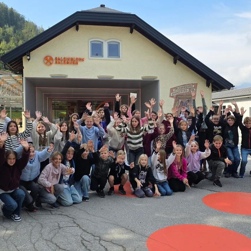 Eine Gruppe von Kindern und Erwachsenen posiert für ein Foto vor einem Gebäude mit einem Schild und Bergen im Hintergrund.