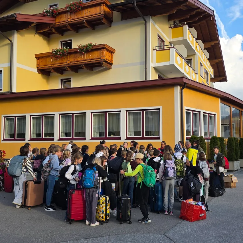 Eine Gruppe von Menschen versammelt sich vor einem gelben Gebäude mit Balkonen, wahrscheinlich auf etwas wartend. Sie tragen Gepäck und einige haben Rucksäcke.