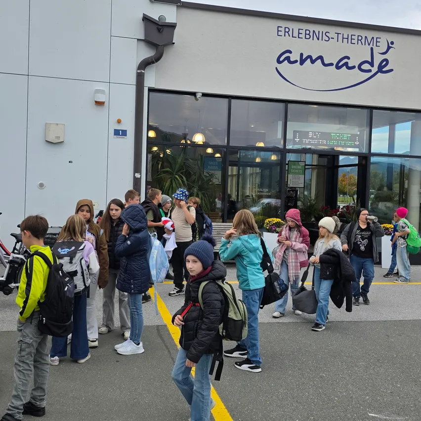 Eine Gruppe von Kindern und Erwachsenen steht vor einem Gebäude mit dem Namen 'Erlebenis-Therme amade'. Sie tragen Rucksäcke und einige halten Taschen.