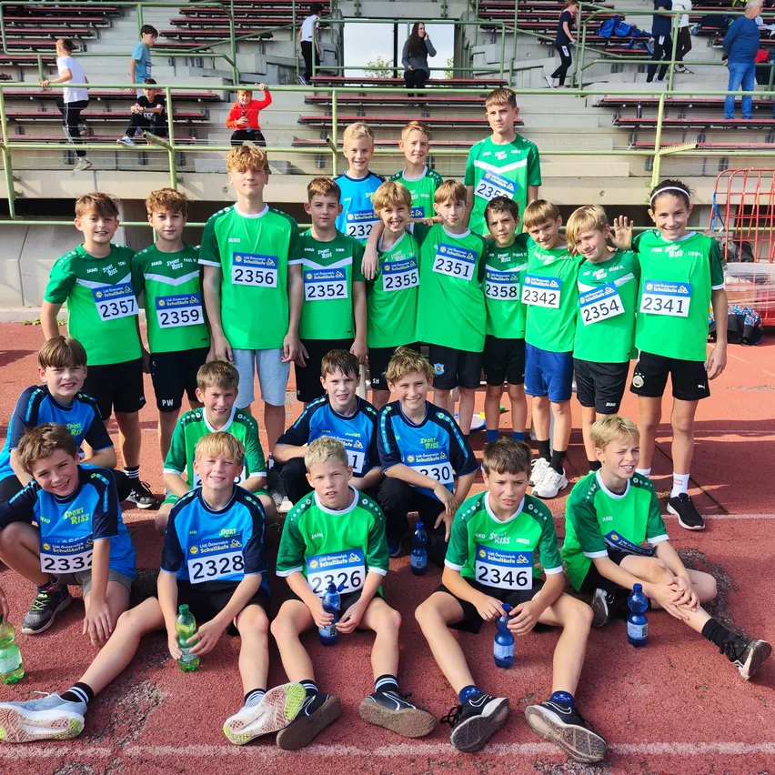 Eine Gruppe junger Athleten in grünen und blauen Uniformen posiert für ein Foto auf einer Laufbahn, mit Nummern auf ihren Trikots und Flaschen in den Händen.