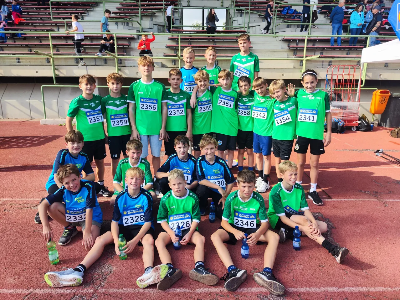 Eine Gruppe junger Athleten in grünen und blauen Uniformen posiert für ein Foto auf einer Laufbahn, mit Nummern auf ihren Trikots und Flaschen in den Händen.