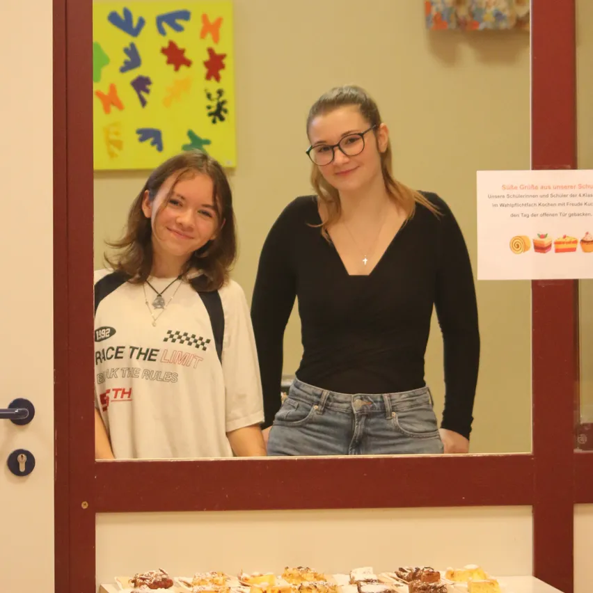 Zwei Frauen stehen hinter einer Tür und lächeln für ein Foto. Eine trägt ein weißes Shirt mit einem Karomuster, die andere ein schwarzes Shirt. Vor ihnen steht ein Tisch mit verschiedenen Desserts.