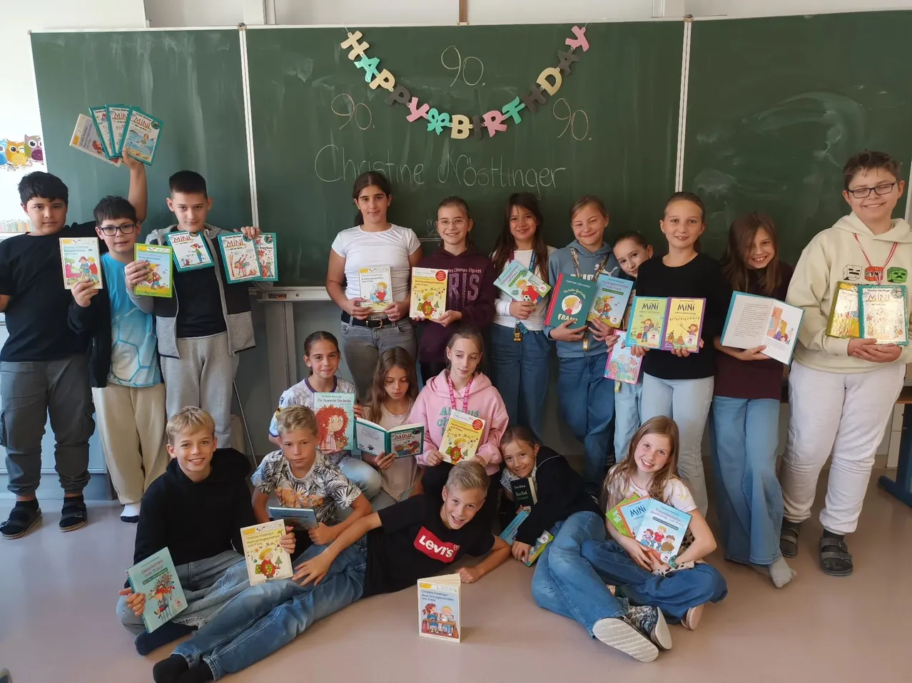 Eine Gruppe von Kindern und Erwachsenen posiert für ein Foto im Klassenzimmer. Sie halten Bücher und lächeln.