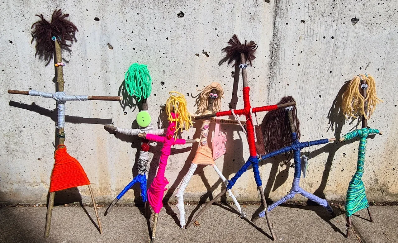 Eine Gruppe von Steckfiguren mit bunten Garnen und Accessoires steht in einer Reihe an einer Betonwand.