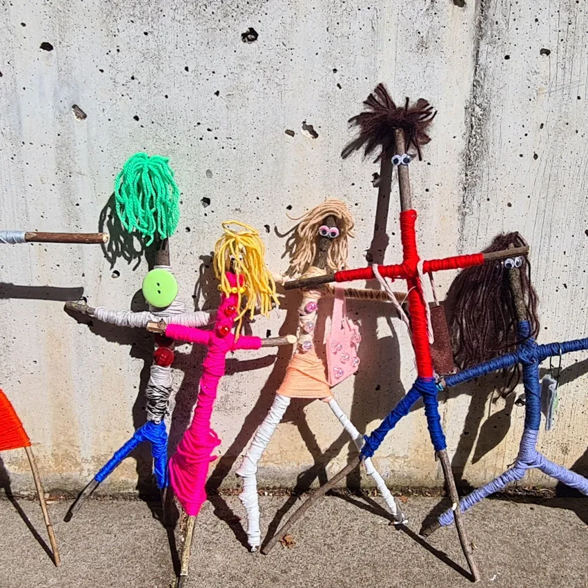 Eine Gruppe von Steckfiguren mit bunten Garnen und Accessoires steht in einer Reihe an einer Betonwand.