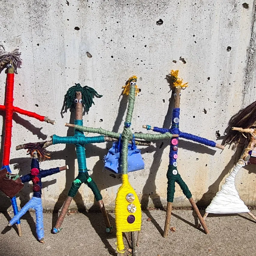 Eine Reihe von Steckfiguren aus Zweigen und Garn steht an einer Betonwand.