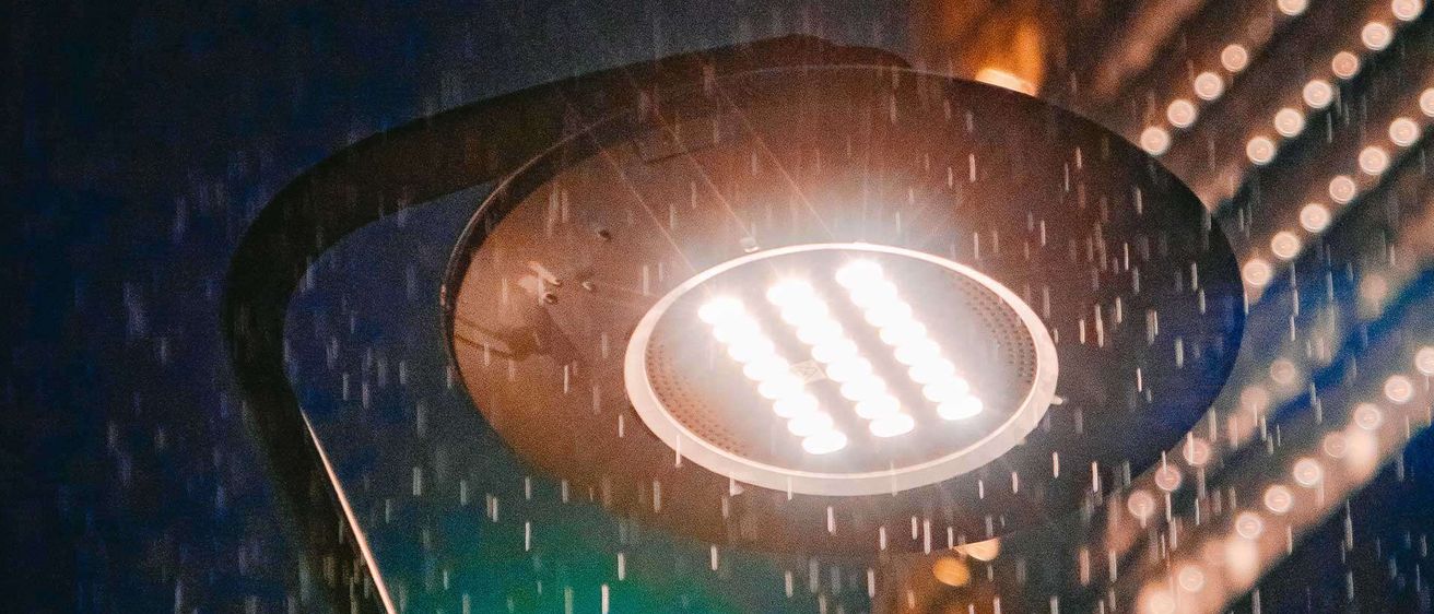 Eine Straßenlaterne mit einem beleuchteten runden Gehäuse strahlt im Regen hell und wirft Licht durch die Regentropfen.