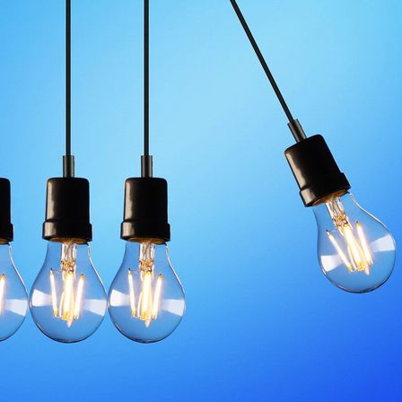 Fünf Glühbirnen hängen an schwarzen Kabeln vor einem blauen Hintergrund. Vier der Glühbirnen leuchten mit einem warmen, gelben Licht.