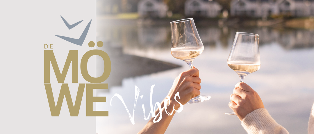 Eine Hand hält ein Weinglas mit 'OE Vibes' vor einem See und Häusern. Das Glas ist mit Weißwein gefüllt.