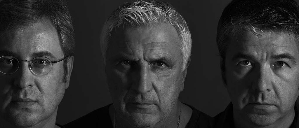 Drei Männer in Schwarz und Weiß posieren für ein Foto. Der Mann in der Mitte trägt eine Halskette.