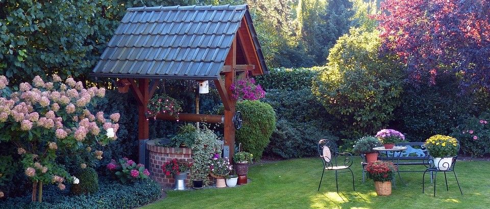Ein hölzerner Pavillon mit einem blauen Dach und einer darunterliegenden Ziegelstruktur, umgeben von verschiedenen Topfpflanzen und Blumen in einem sonnigen Garten.