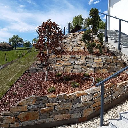 Eine Steinmauer mit Stufen, umgeben von Kies und einem kleinen Baum, führt zu einem Haus unter einem blauen Himmel mit Wolken.
