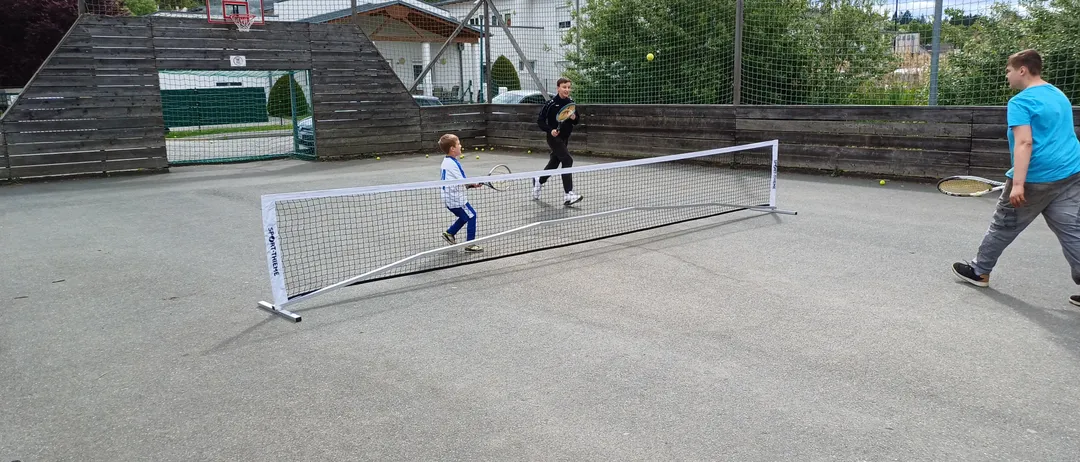 Zwei Kinder spielen Tennis auf einem Platz mit Netz. Ein Junge bereitet sich darauf vor, den Ball zu schlagen. Der Platz ist von einem Zaun umgeben und hat einen Basketballkorb.