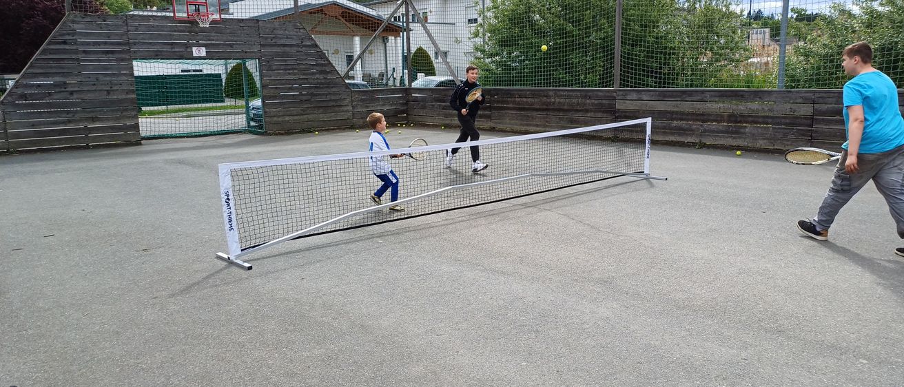 Zwei Kinder spielen Tennis auf einem Platz mit Netz. Ein Junge bereitet sich darauf vor, den Ball zu schlagen. Der Platz ist von einem Zaun umgeben und hat einen Basketballkorb.