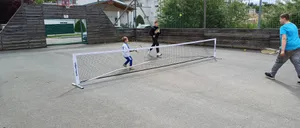 Zwei Kinder spielen Tennis auf einem Platz mit Netz. Ein Junge bereitet sich darauf vor, den Ball zu schlagen. Der Platz ist von einem Zaun umgeben und hat einen Basketballkorb.