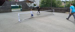 Zwei Kinder spielen Tennis auf einem Platz mit Netz. Ein Junge bereitet sich darauf vor, den Ball zu schlagen. Der Platz ist von einem Zaun umgeben und hat einen Basketballkorb.