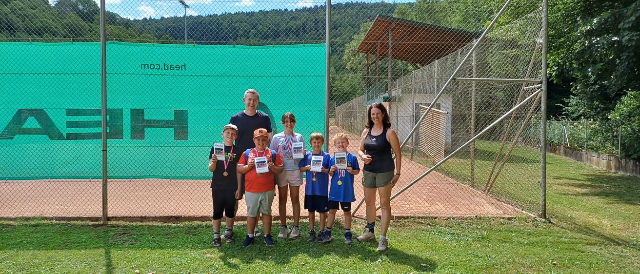 Eine Gruppe junger Athleten und ihrer Trainer steht auf einem Rasenfeld, hält Medaillen und Urkunden in der Hand, mit einem Tennisplatz und einem grünen Netz im Hintergrund.